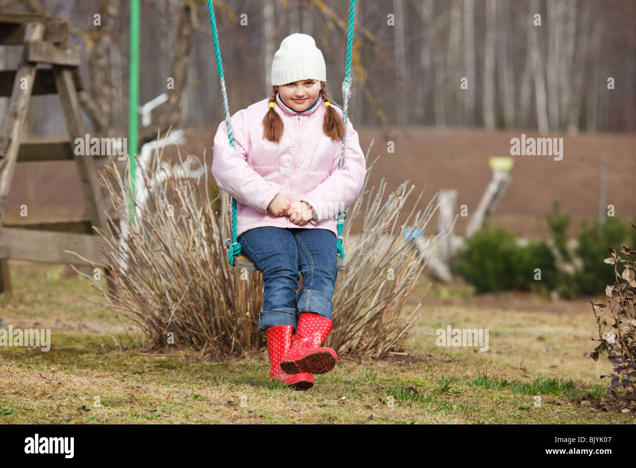 Little girl sitting in swings in a garden. Early spring with first sunbeams. Stock Photo