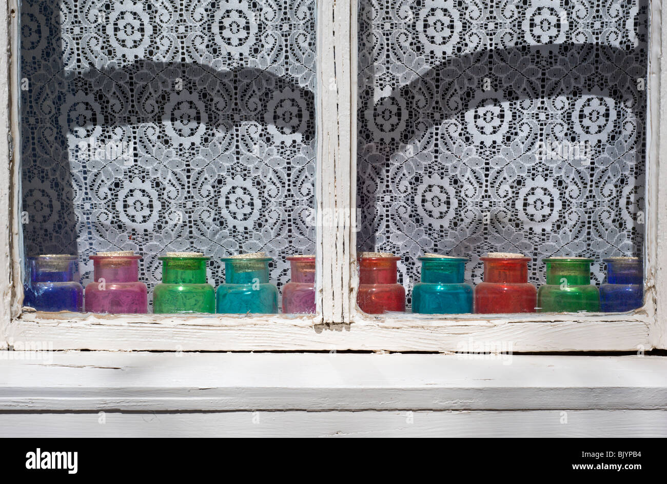 An old white-washed window, with lace curtains, has red, green and blue glass jars sitting on the sill, in Lincoln, New Mexico. Stock Photo