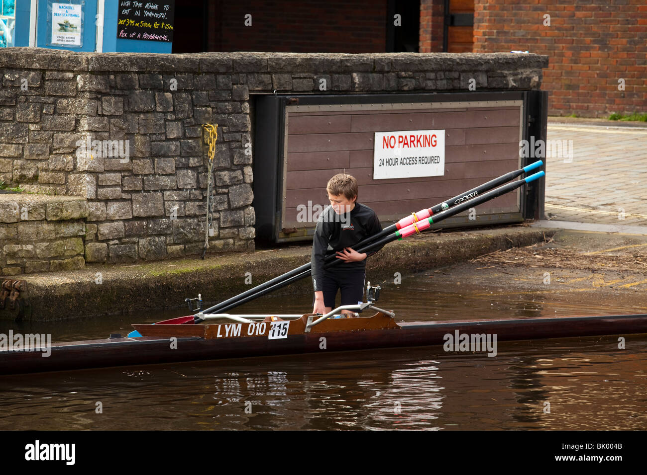 Teenage Rower teenage boy in wetsuit holding oars launching single skull rowing boat ...