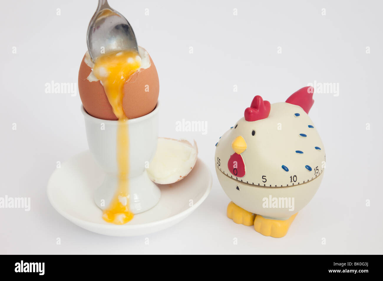 Spoon in an open soft boiled egg with runny yolk in an egg cup with a chicken egg timer on a white background Stock Photo