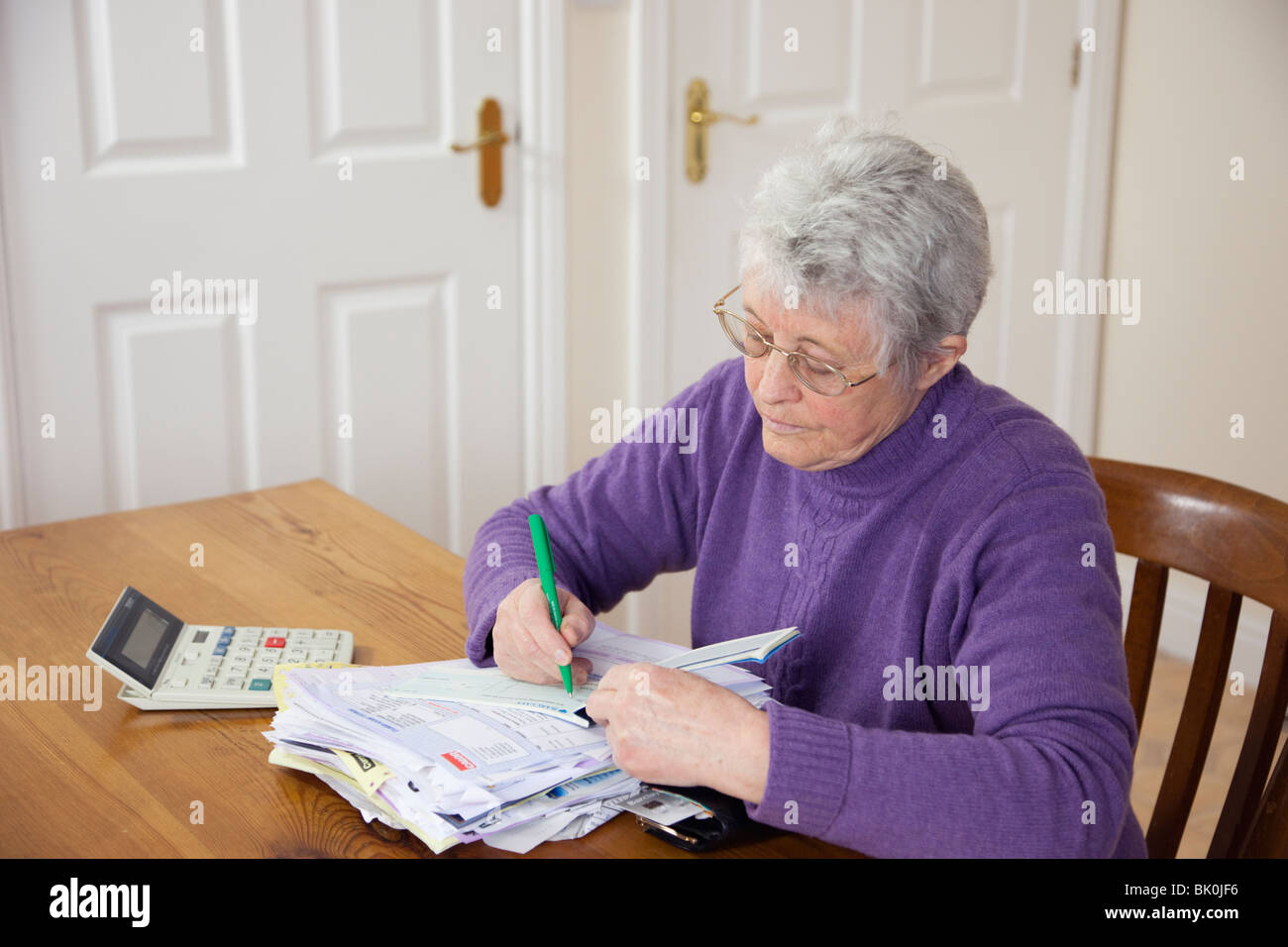 Senior woman pensioner with a big pile of bills on the table writing a cheque to pay a bill at home. England, UK, Britain. Stock Photo