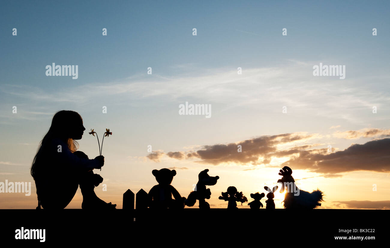 Young girl with daffodils and a Rag doll, chicken, fox, rabbit and bear soft toys sitting on a gate at sunset . Silhouette Stock Photo