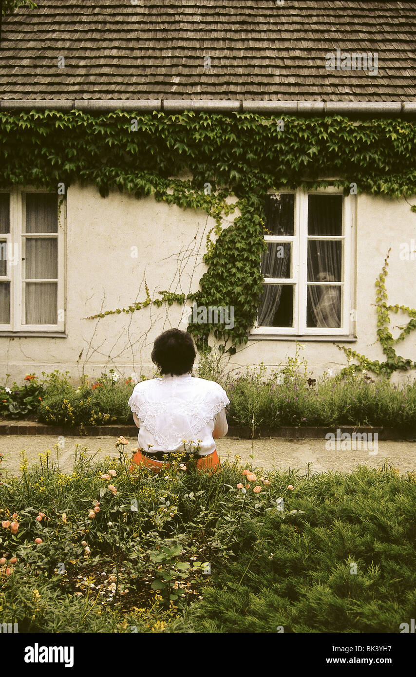 Woman sitting by a garden with a house with vines and windows in the background, Poland Stock Photo