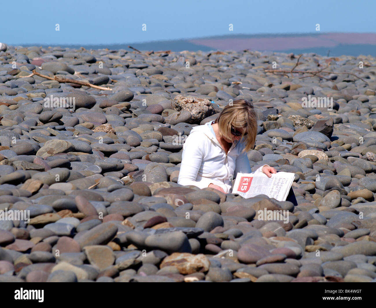 Woman sat in amongst the pebbles at the beach reading the paper, Westward Ho!, Devon Stock Photo