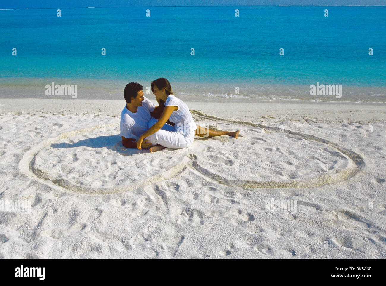 Young couple on beach sitting in a heart shaped imprint on the sand, Maldives, Indian Ocean, Asia Stock Photo