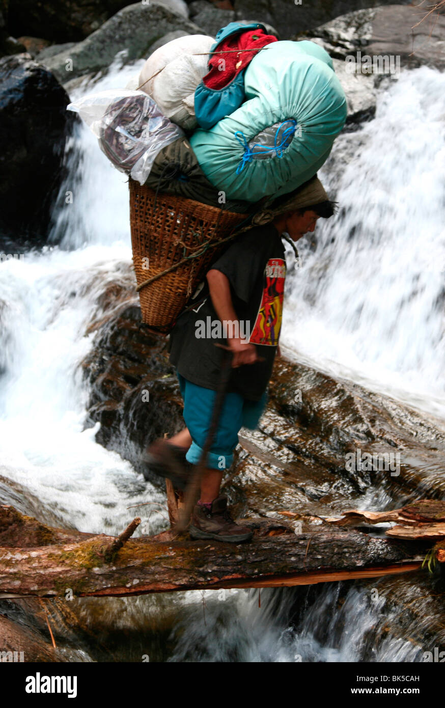 A porter crosses a river carrying a heavy load up the Hingku Valley, near Lukla, south of Mount Everest, Nepal Stock Photo