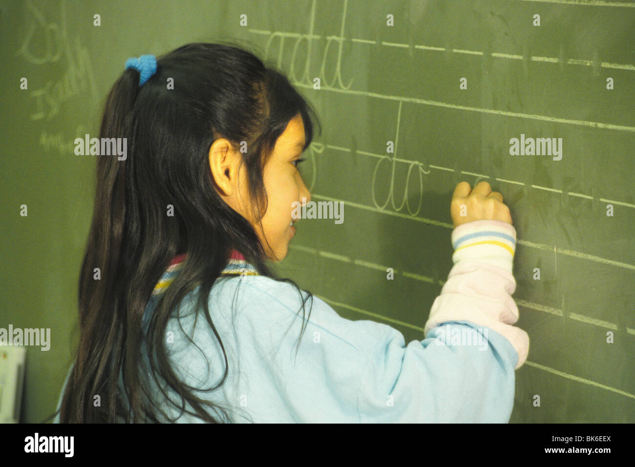 released ethnic girl elementary school student practices handwriting on blackboard Stock Photo
