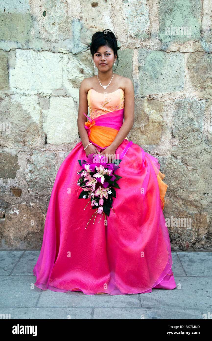 pretty dark eyed Mexican teen with upswept hair dressed in pink organza gown & carrying  pink orchids for her Quinceanera Stock Photo