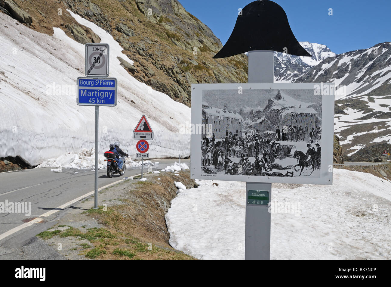 Sign depicting Napoleon's crossing of Great St Bernard Pass with Motorbike starting descent past snow drifts towards Martigny Stock Photo