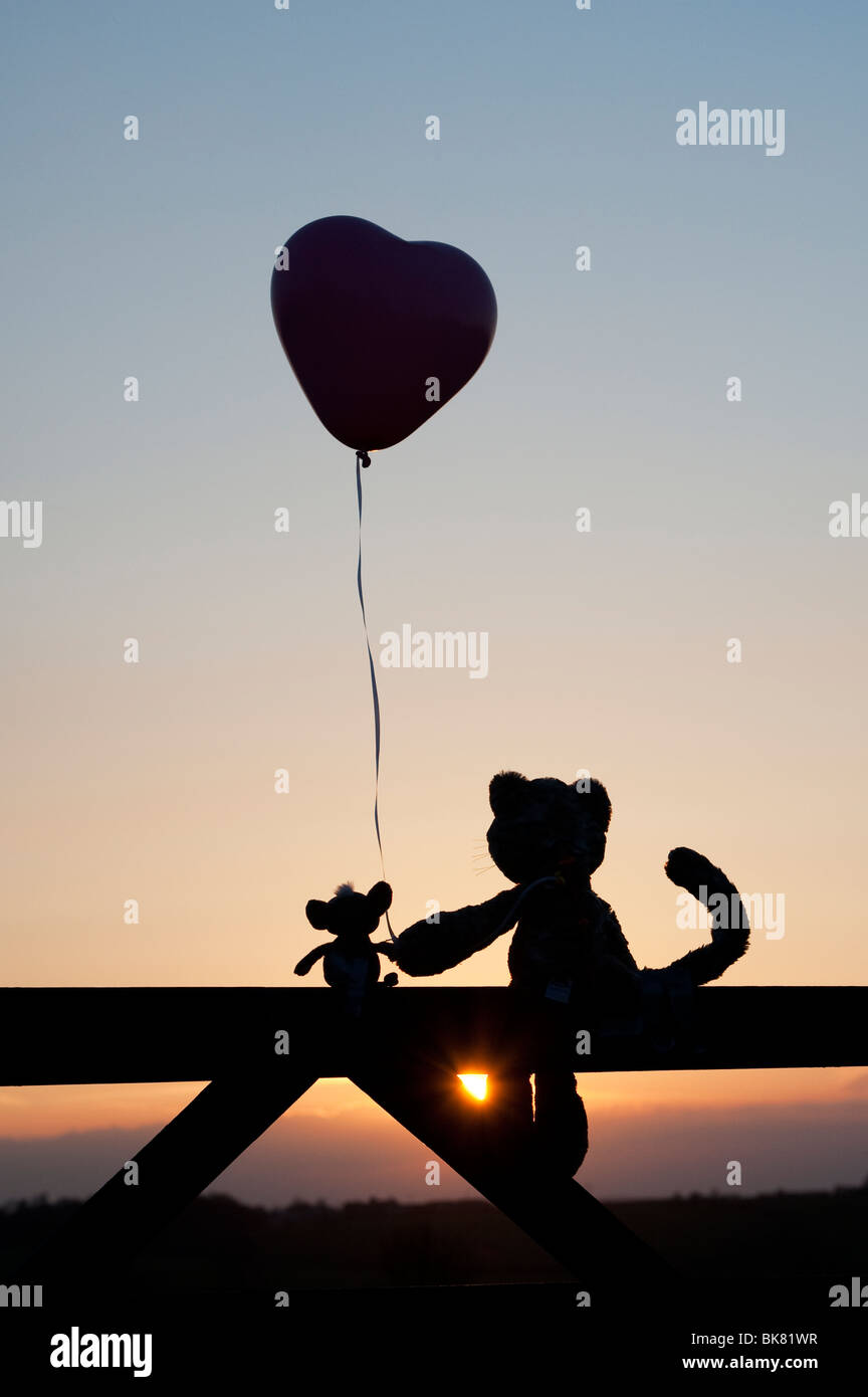 Cat and mouse soft toys holding a heart shape balloon in friendship, sitting on a gate at sunset. Silhouette Stock Photo