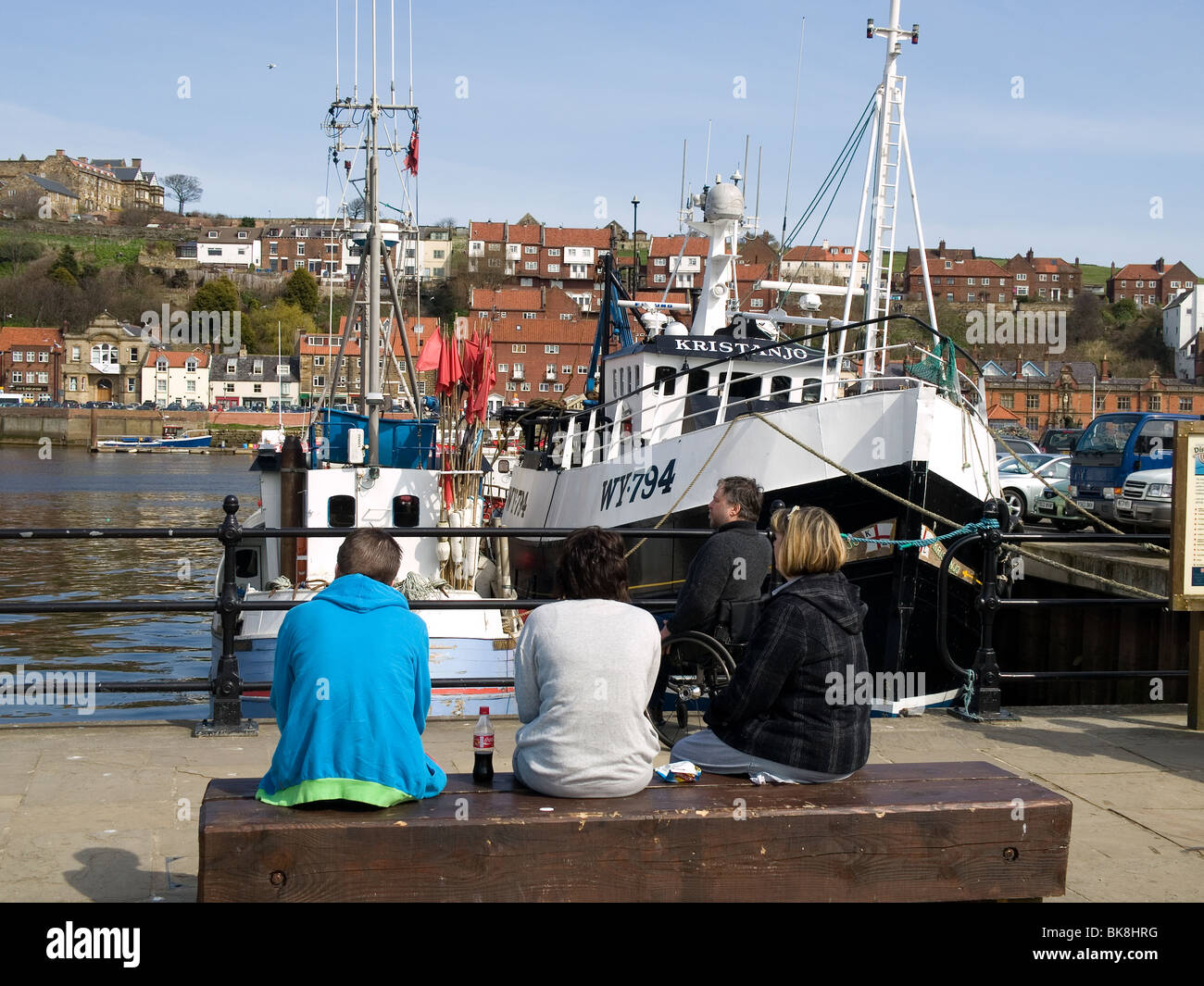 A group of sightseers one in a wheelchair sit and relax in the sunshine looking at the fishing boats at Endeavour  Wharf Whitby Stock Photo
