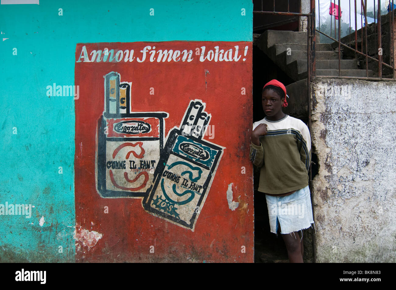 A man stands next to a painted wall advertising local brand cigarette company in Port au Prince Haiti Stock Photo