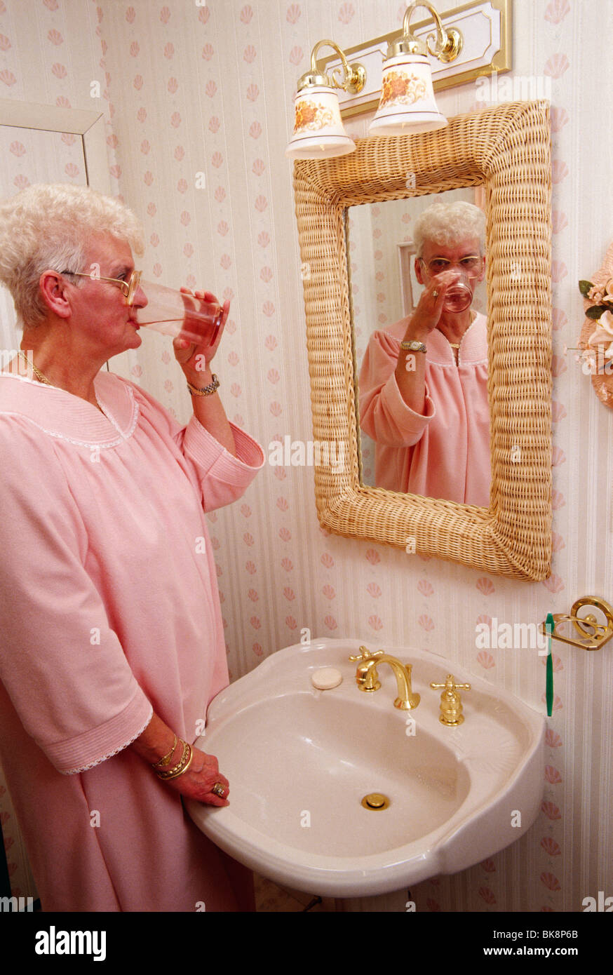 Elderly woman in her bathrobe drinking fresh water from a glass in her bathroom. Stock Photo