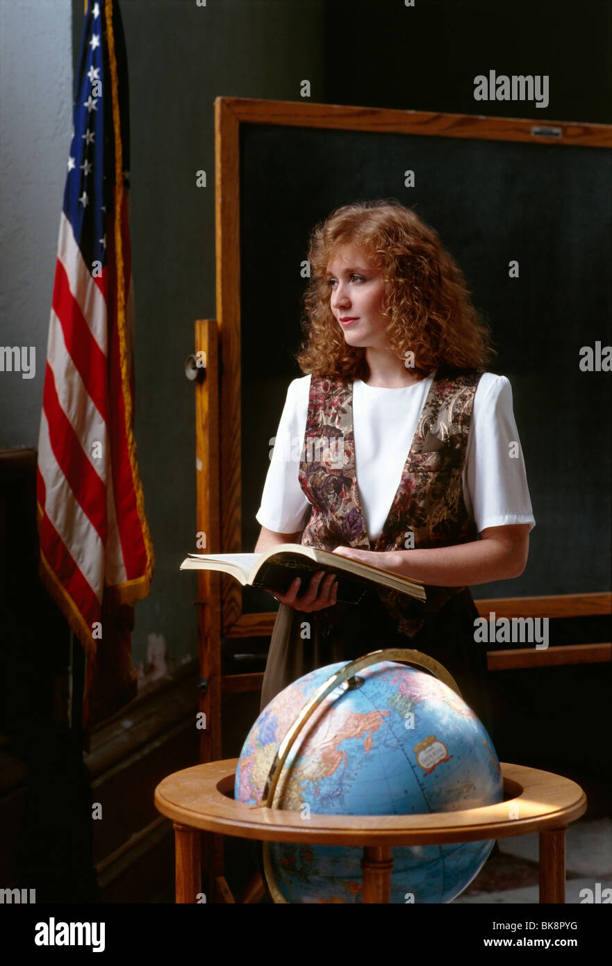 Female teacher with open book, world globe, American Flag, and blackboard in a classroom. Stock Photo