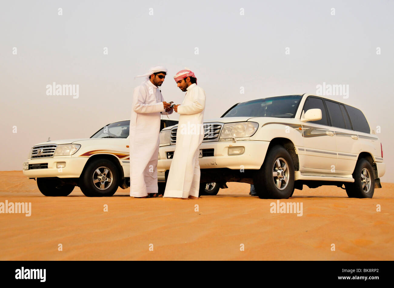 Two Arabs in Dishdashas, the typical white cloaks, with jeeps and mobile phones in the desert, Liwa Oasis, Abu Dhabi, United Ar Stock Photo