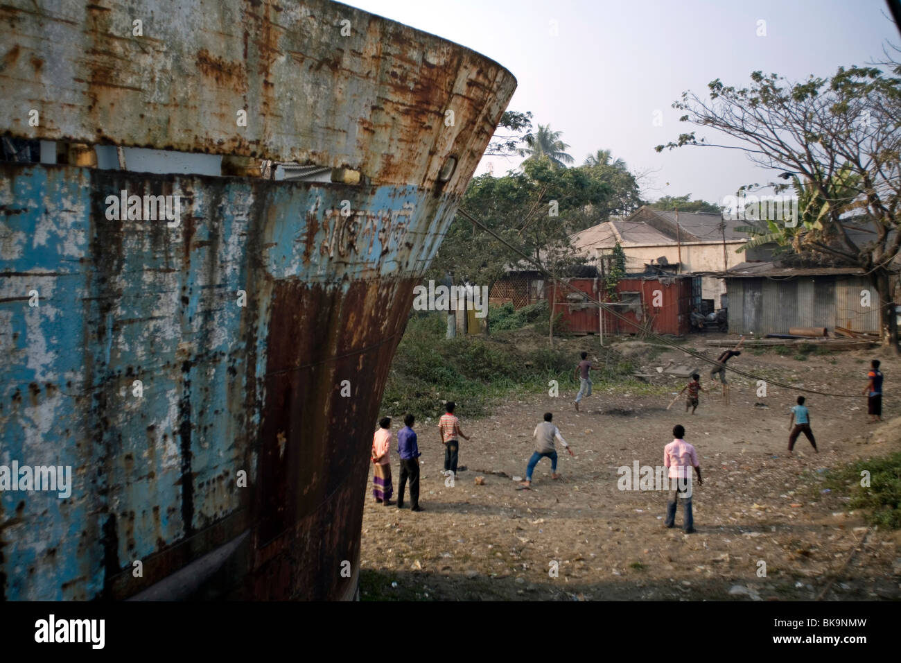 Boys play cricket in the shadow of a rusty hull of a ship in the docks at Sadarghat, Chittagong, Bangladesh Stock Photo