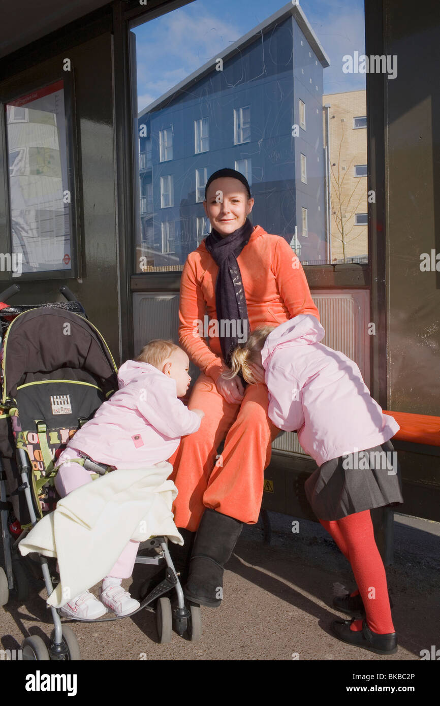 A mum in an orange tracksuit waits for the bus with her two daughters. Stock Photo