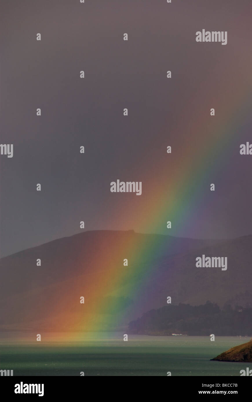 A rainbow in passing rain showers over the sea with coastal hills in the background Stock Photo