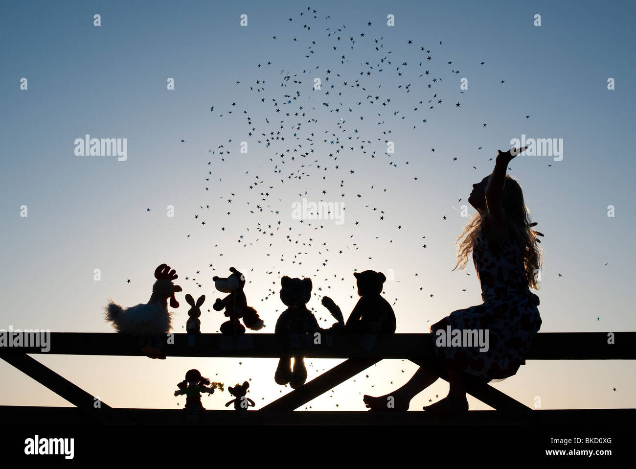 Girl throwing stars over a Rag doll, chicken, fox, rabbit and teddy bear soft toys sitting on a gate at sunset. Silhouette Stock Photo