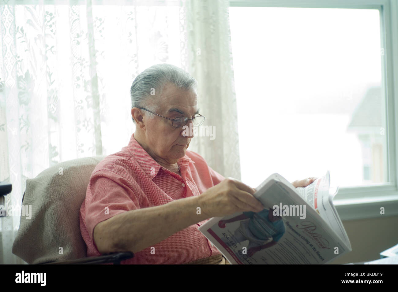 Old man sits in living room chair by window, reading newspaper Stock