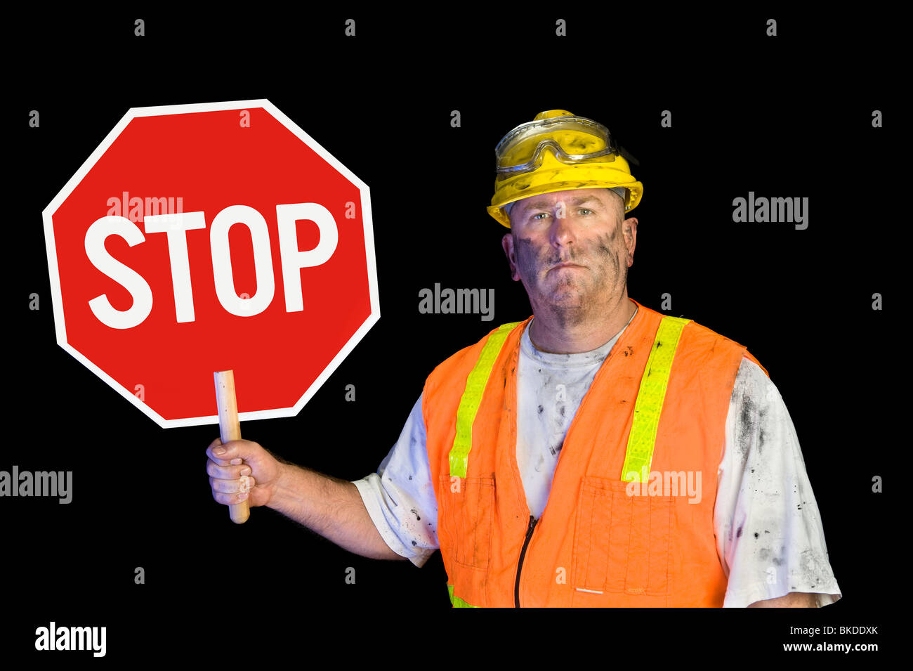 A dirty, grungy, greasy utility construction worker with hard hat, orange vest and eye protection holds up a stop sign. Stock Photo