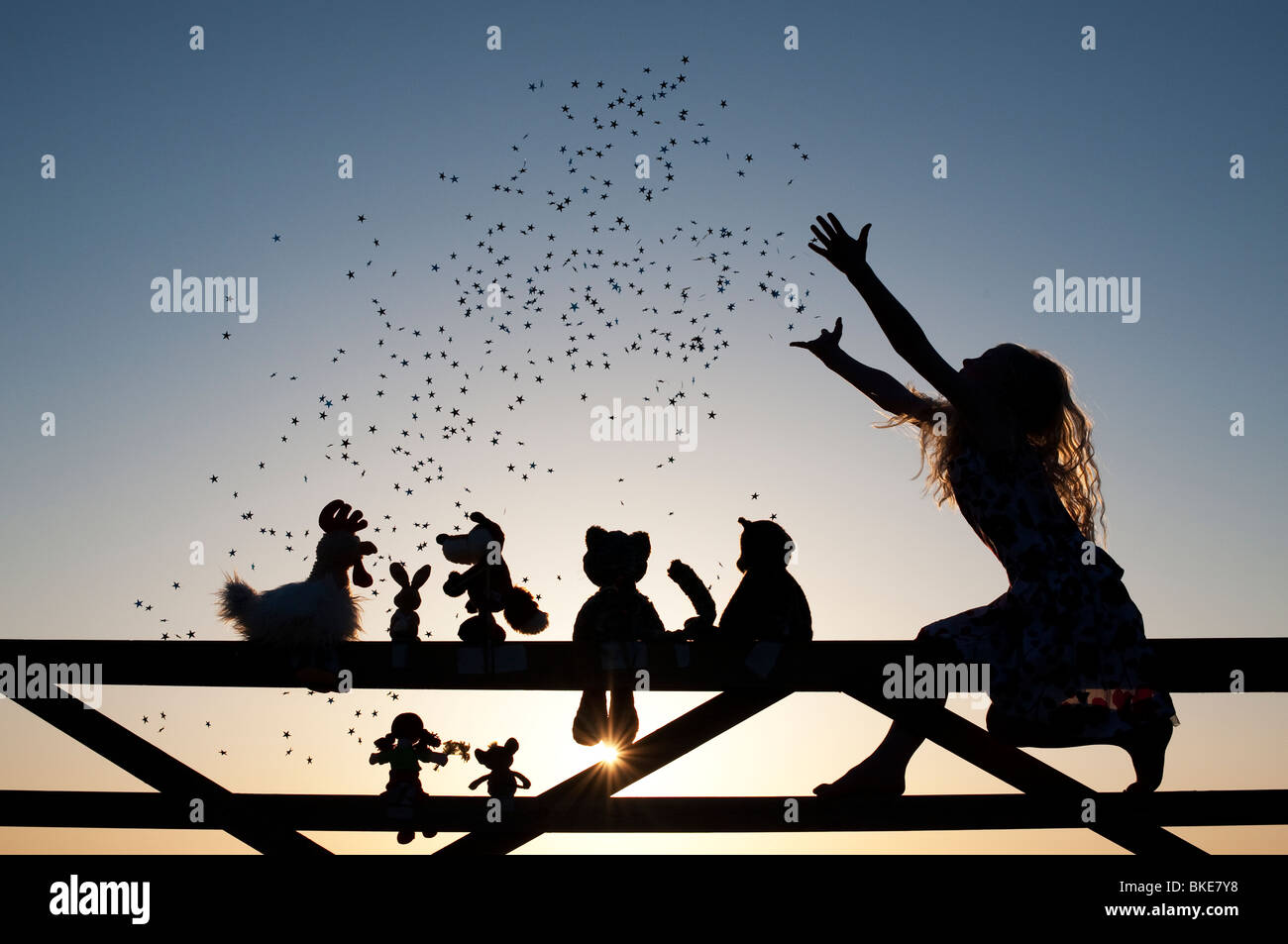 Girl throwing stars over a Rag doll, chicken, fox, rabbit, cat and teddy bear soft toys sitting on a gate at sunset. Silhouette Stock Photo