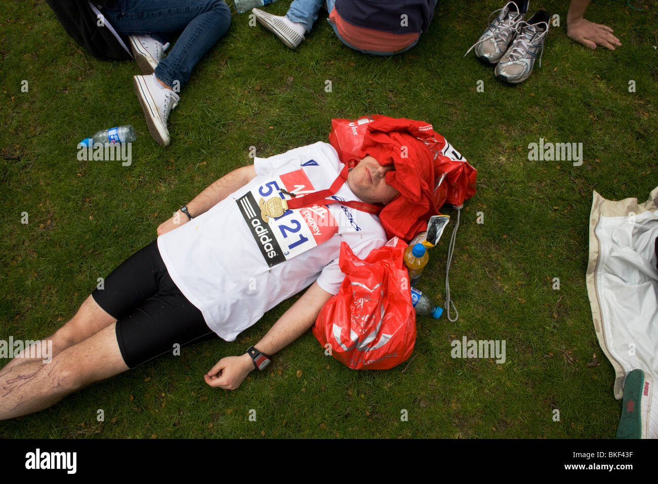 With face covered and medal around neck, a London Marathon runner collapsed on grass, before being met by family Stock Photo