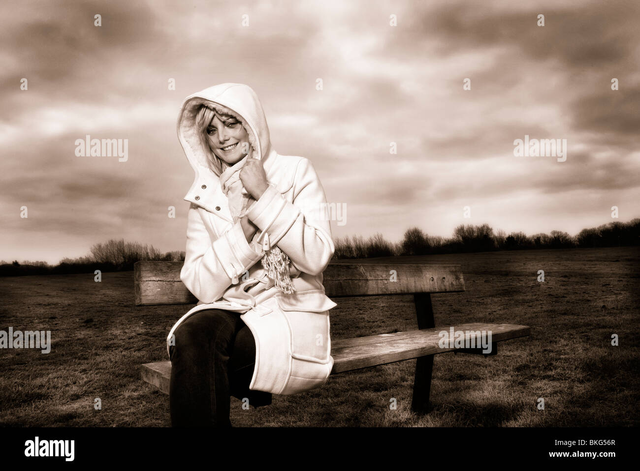 blonde woman sitting on park bench smiling with hood up Stock Photo