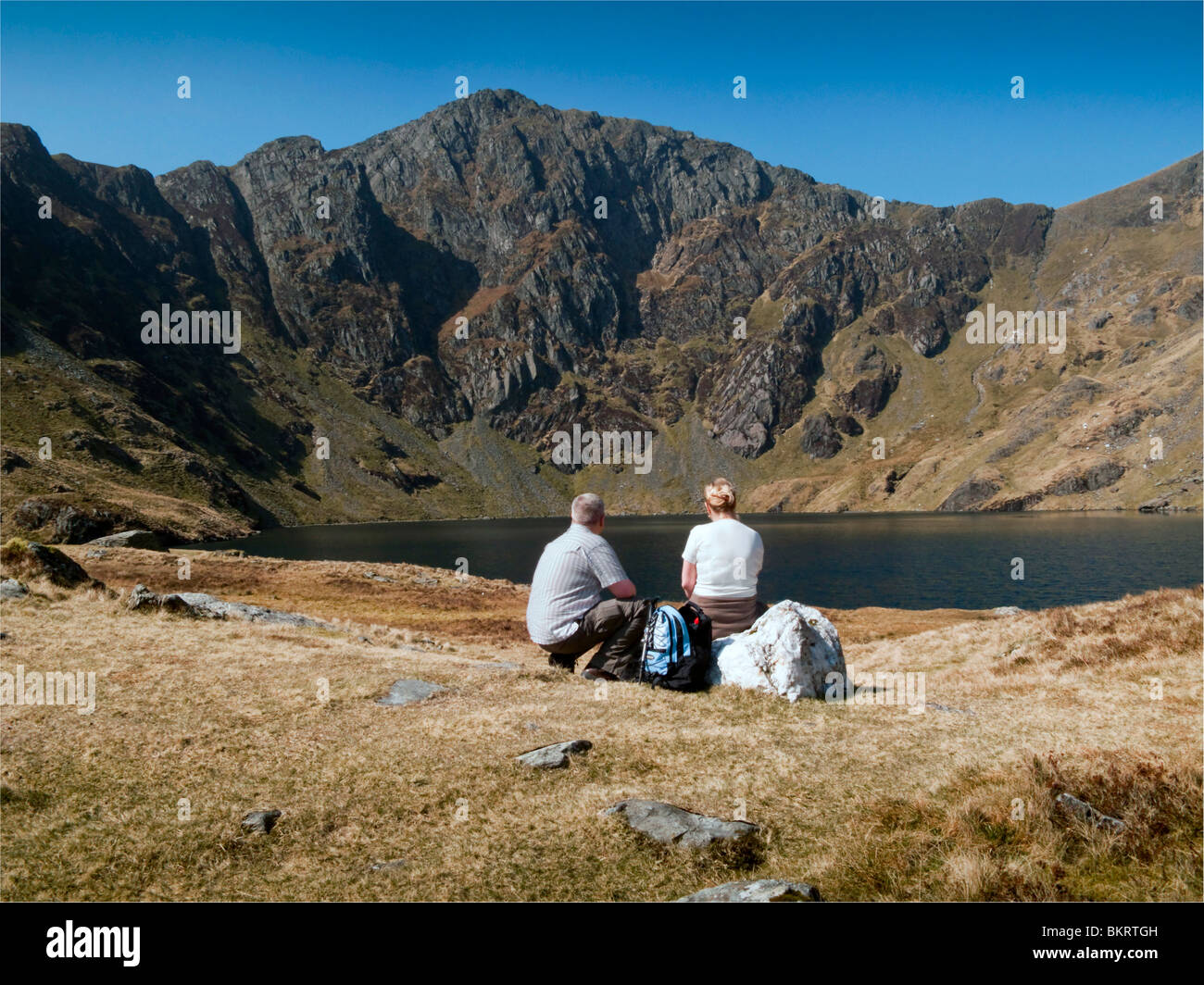 Mature walkers male and female relaxing infront of Lake Cau looking up at  Craig Cau with backpacks in Snowdonia national park Stock Photo