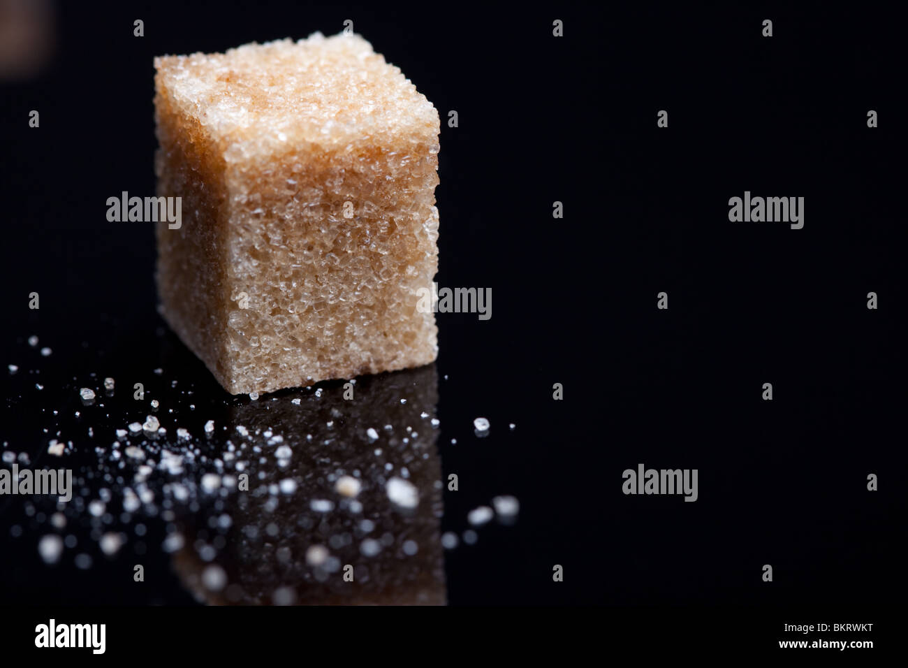 A single brown sugar cube and some sugar granules on a black reflective surface Stock Photo