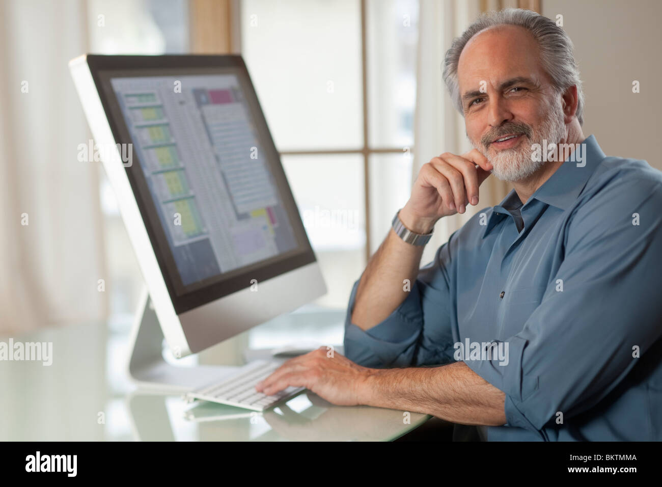 Portrait of a businessman dressed in casual clothing and sitting in front of a computer. He is smiling at the camera. Stock Photo