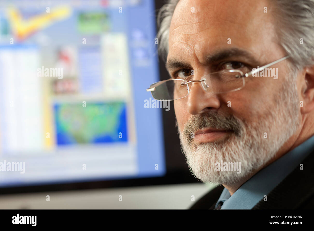 Close-up portrait of a businessman wearing eyeglasses and looking at the camera with a computer monitor in the background. Stock Photo