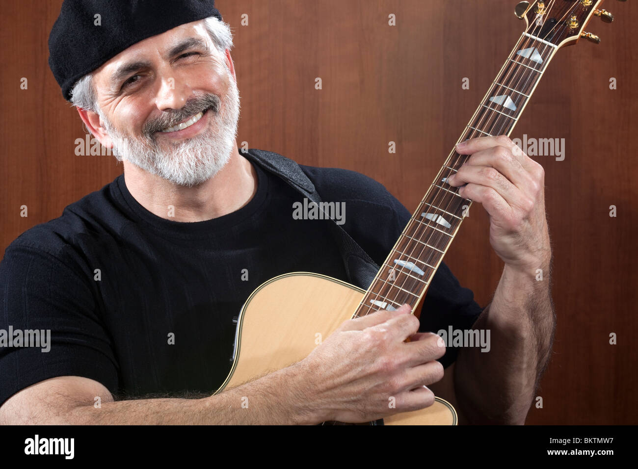 Portrait of a middle-aged man wearing a black beret and t-shirt and playing an acoustic guitar. He is smiling at the camera. Stock Photo