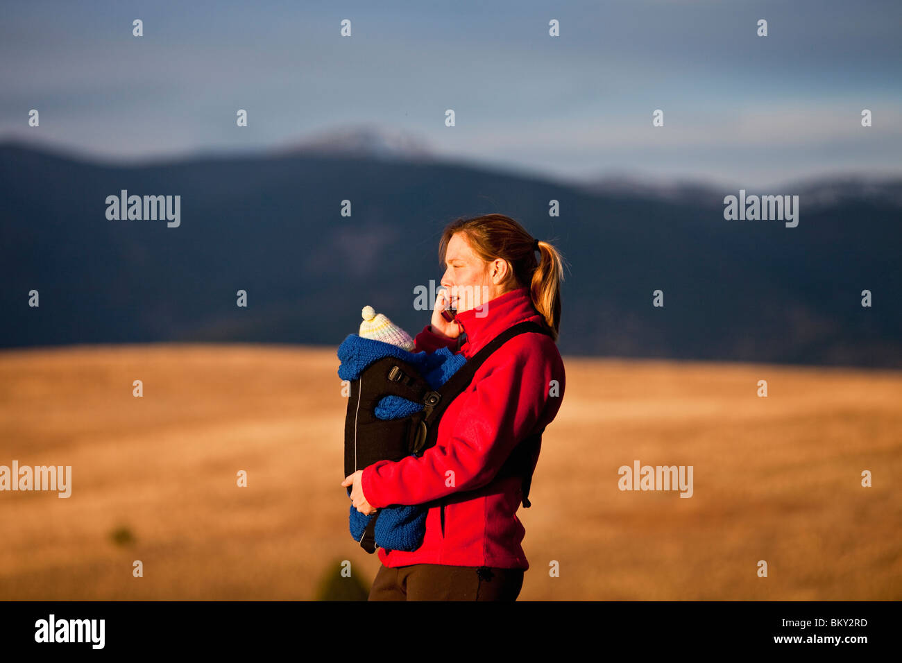 Mother on cell phone and newborn baby in front carrier while hiking through grassy hills, Water