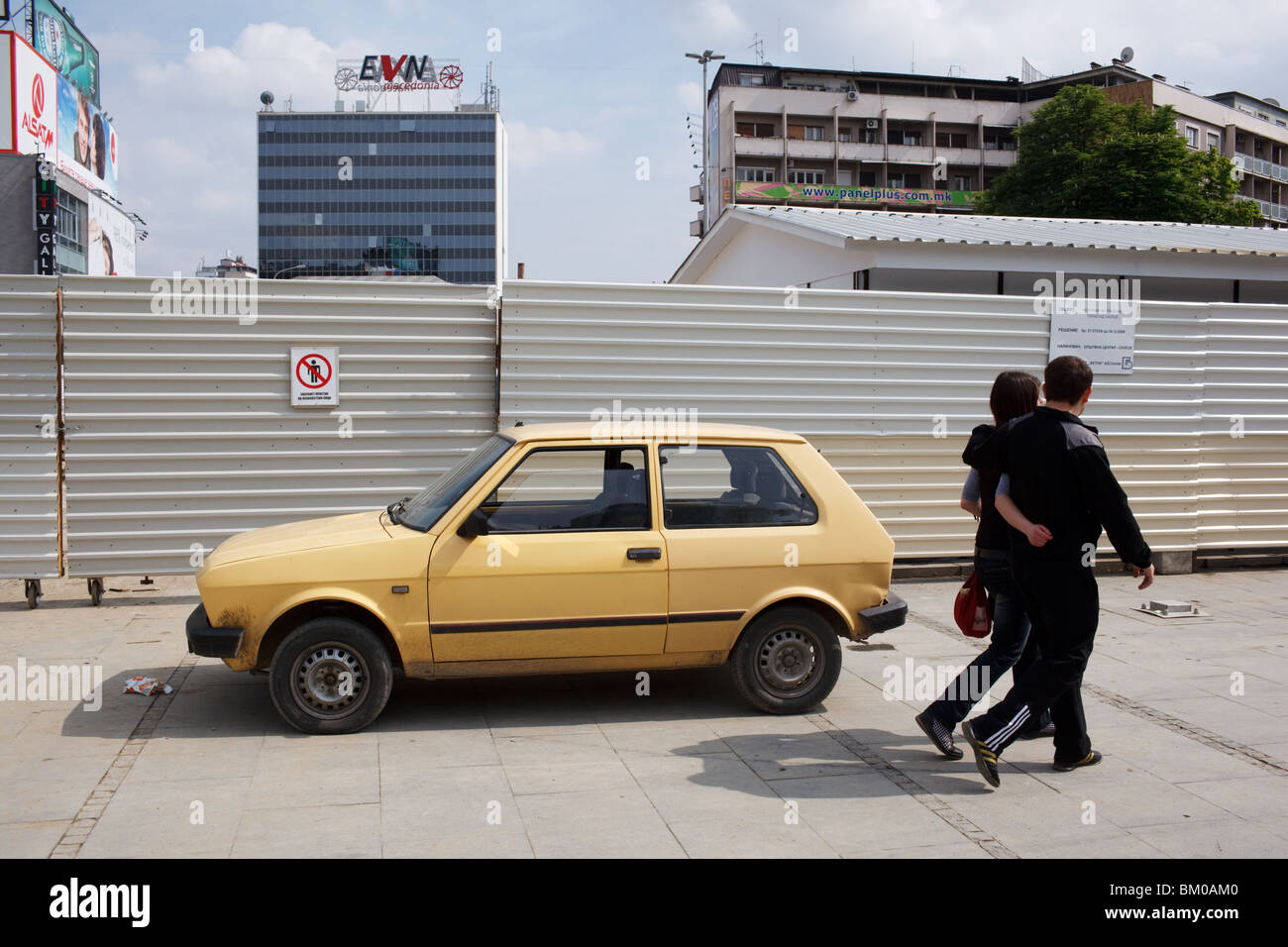 A young couple walks by a yellow Yugo car parked in central Skopje, Macedonia. Stock Photo