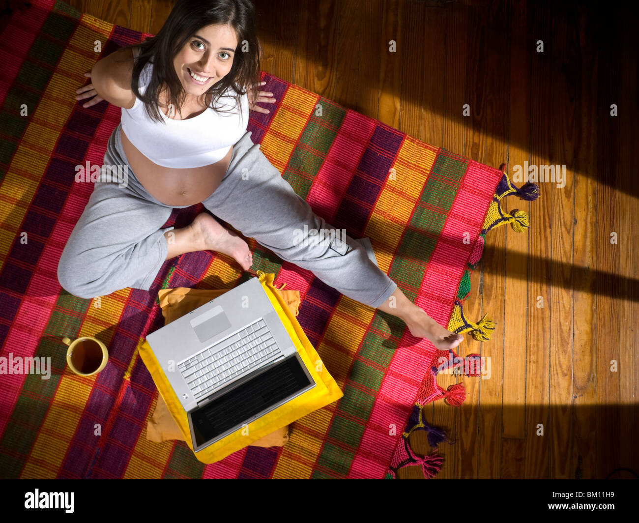 Top view of a young pregnant woman seated on a colorful rug with a laptop computer and a cup of tea. Stock Photo