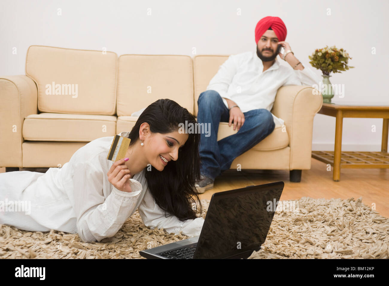 Woman holding a credit card and using a laptop and her husband sitting on couch Stock Photo