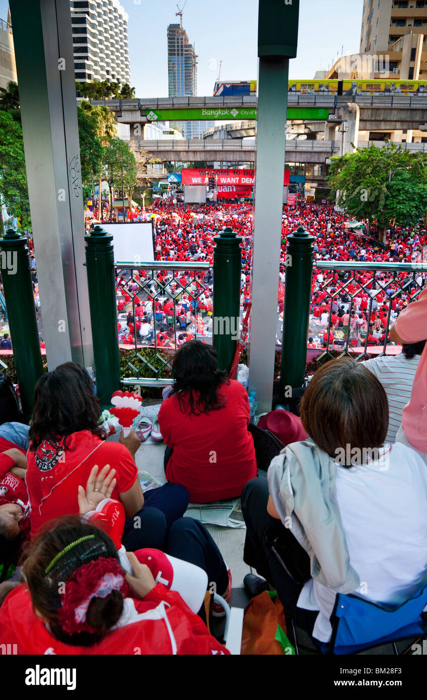 Crowds of red-shirted protesters listen to speeches. A sky-train travels on the track above a stage  with a banner for democracy Stock Photo