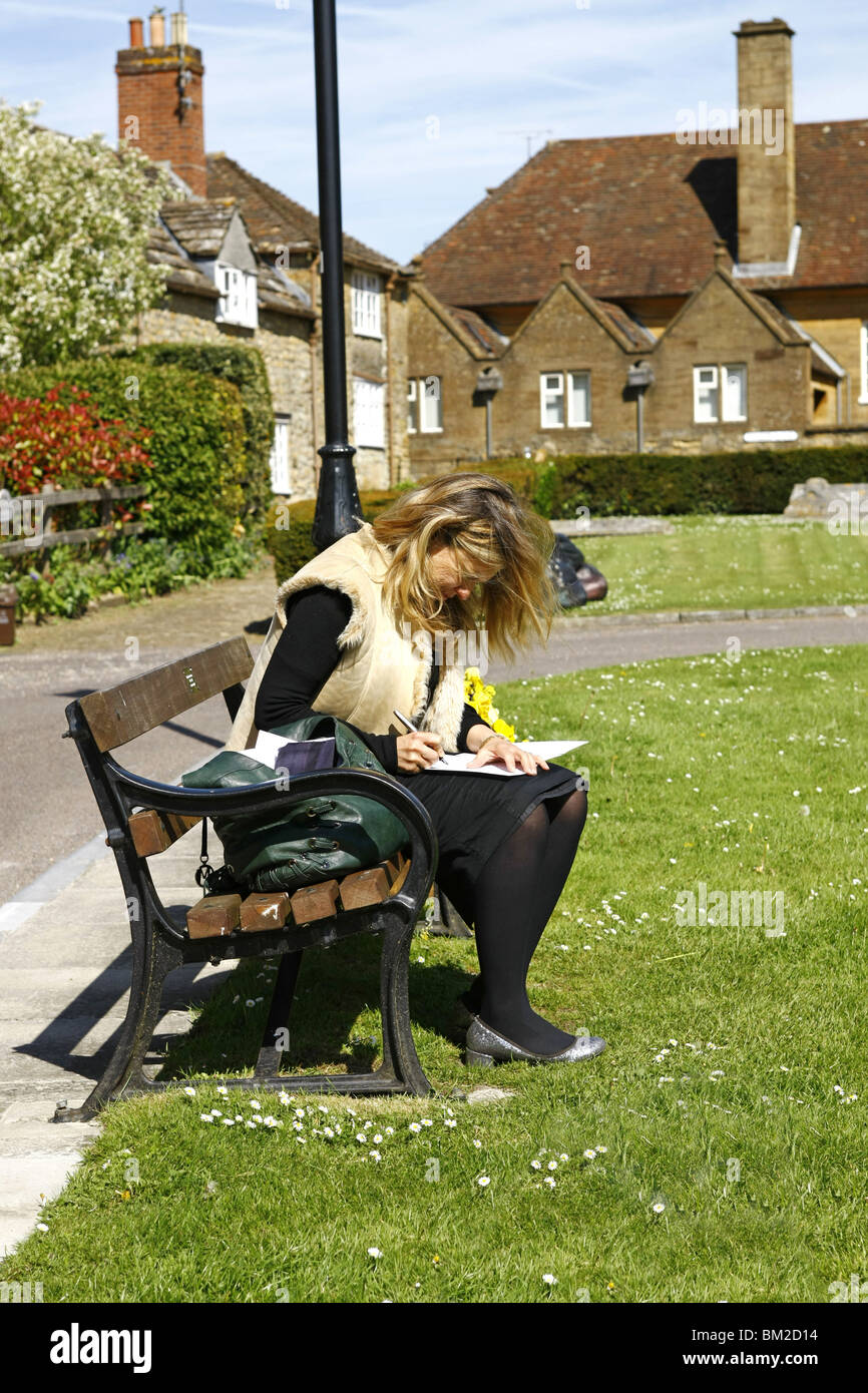 A young woman sits on a park bench in the sunshine writing a message in a Get Well Soon card to her friend. Stock Photo