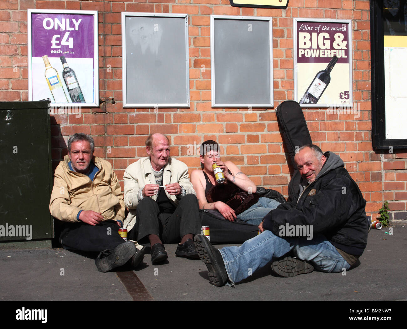 Homeless people drinking alcohol beneath posters advertising cheap alcohol in a U.K. city. Stock Photo