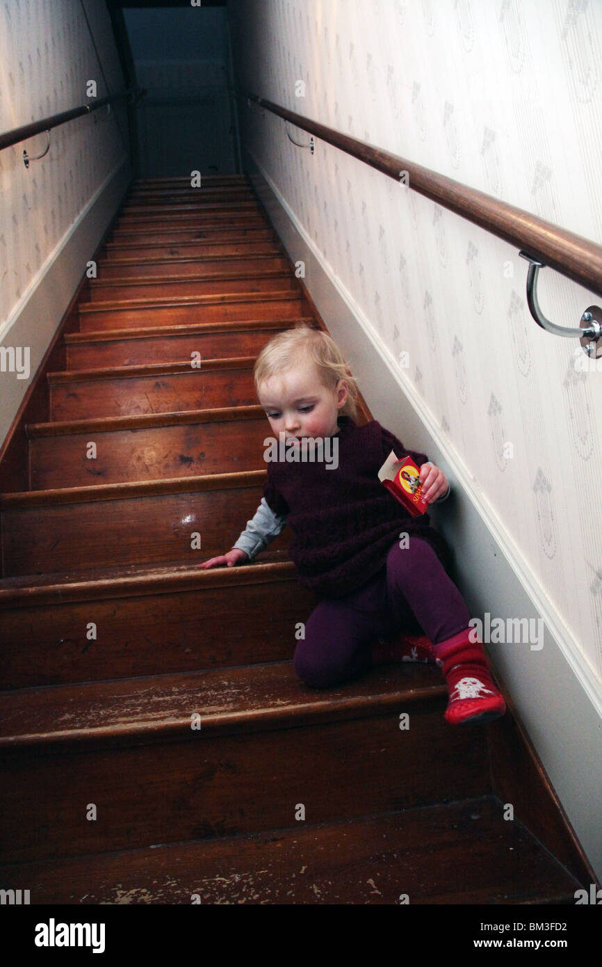 TODDLER, CLIMBING, STAIRS: A two year old baby girl climbs down steep stairs wooden floorboards steps alone box sweets sock boots model released Stock Photo