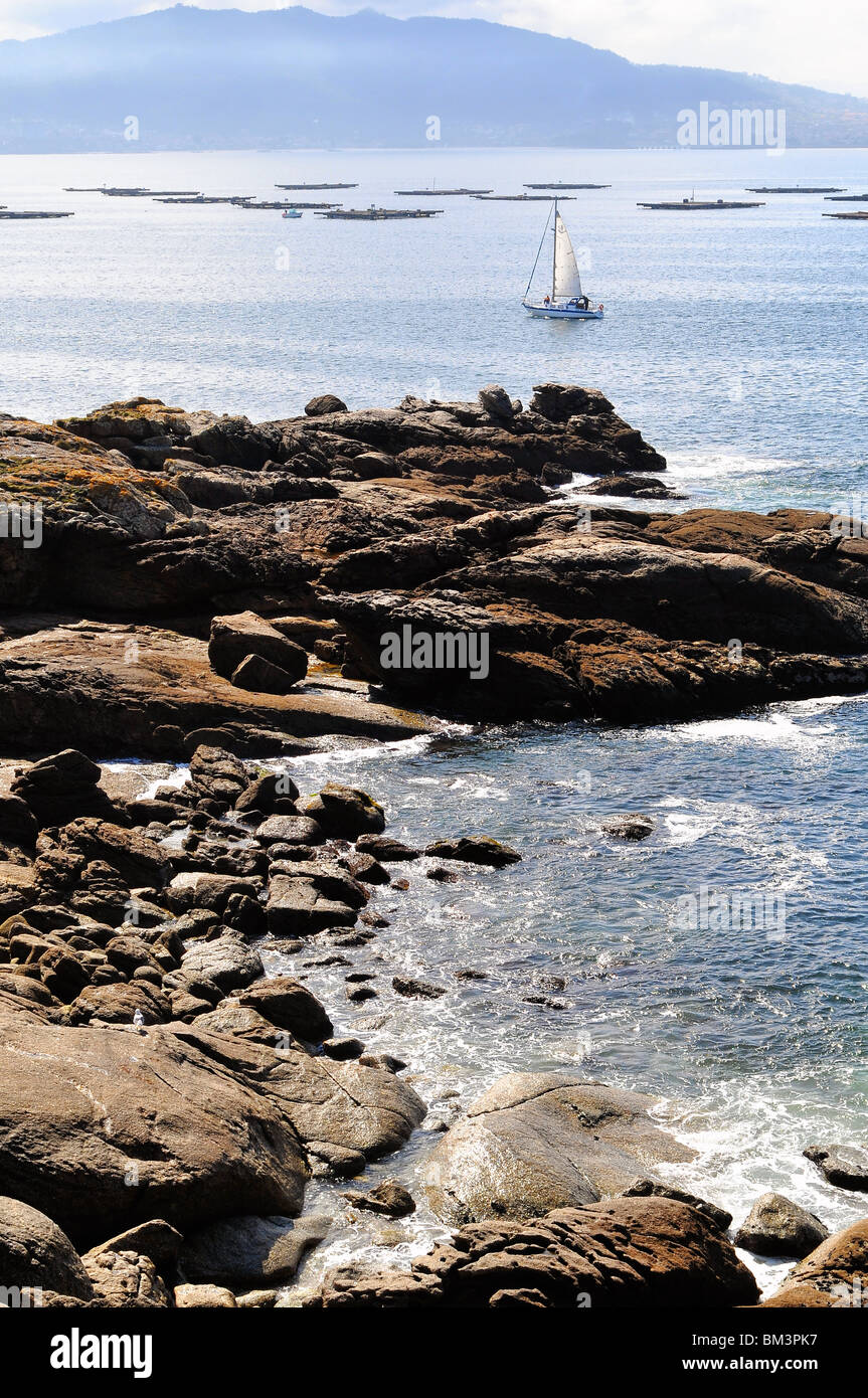 Rock Shore of Galicia Coast, Sailboat sailing on Rias Baixas, Atlantic Ocean, Mussels Rafts and Vigo on a misty background. Stock Photo
