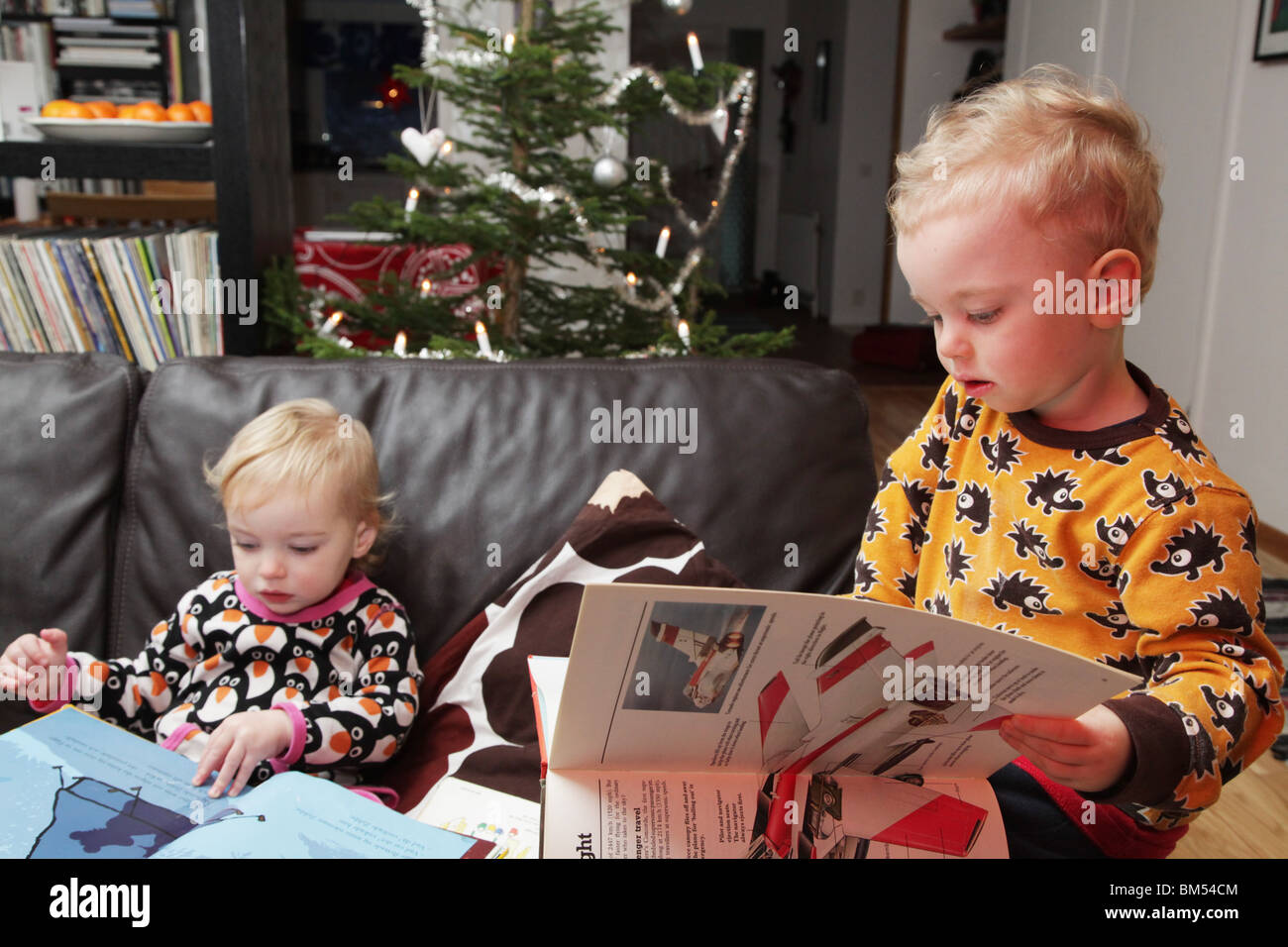 A brother and sister baby toddlers sit at home together reading books next to a Christmas Tree Stock Photo