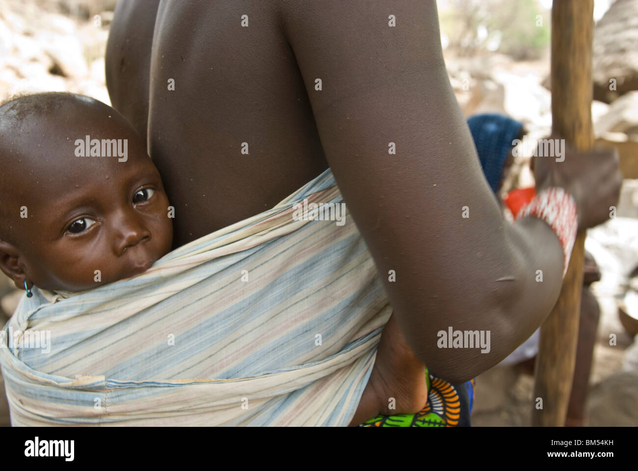 African bedik woman carrying a child while doing housework, Iwol village, Bassari country, Senegal, Africa. Stock Photo