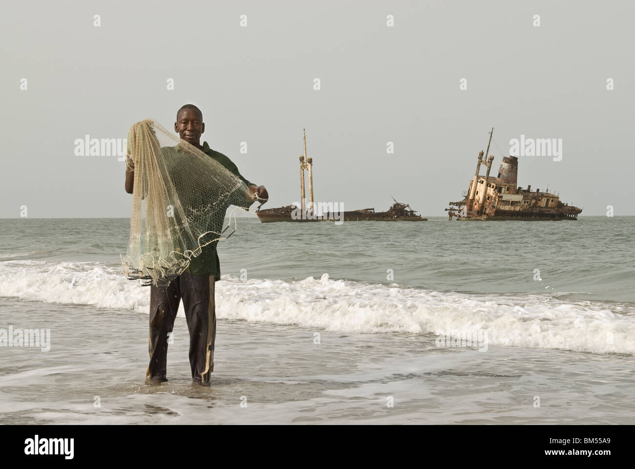 Man fishing with a net from the shore in front of a shipwreck, Palmarin, Senegal, Africa. Stock Photo