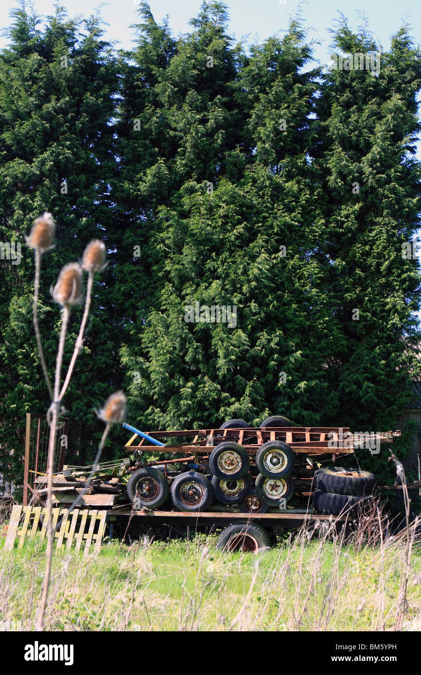 a broken trailer with rusty old trailers stacked on top, in front of conifers in the countryside on a sunny day Stock Photo