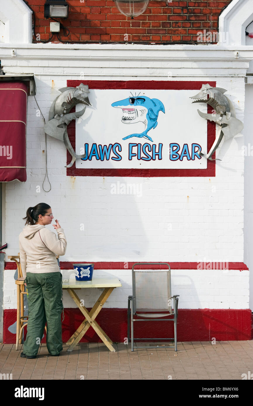 A female adult smoker below a fish bar sign depicting a shark cartoon character on Brighton front Sussex UK Stock Photo