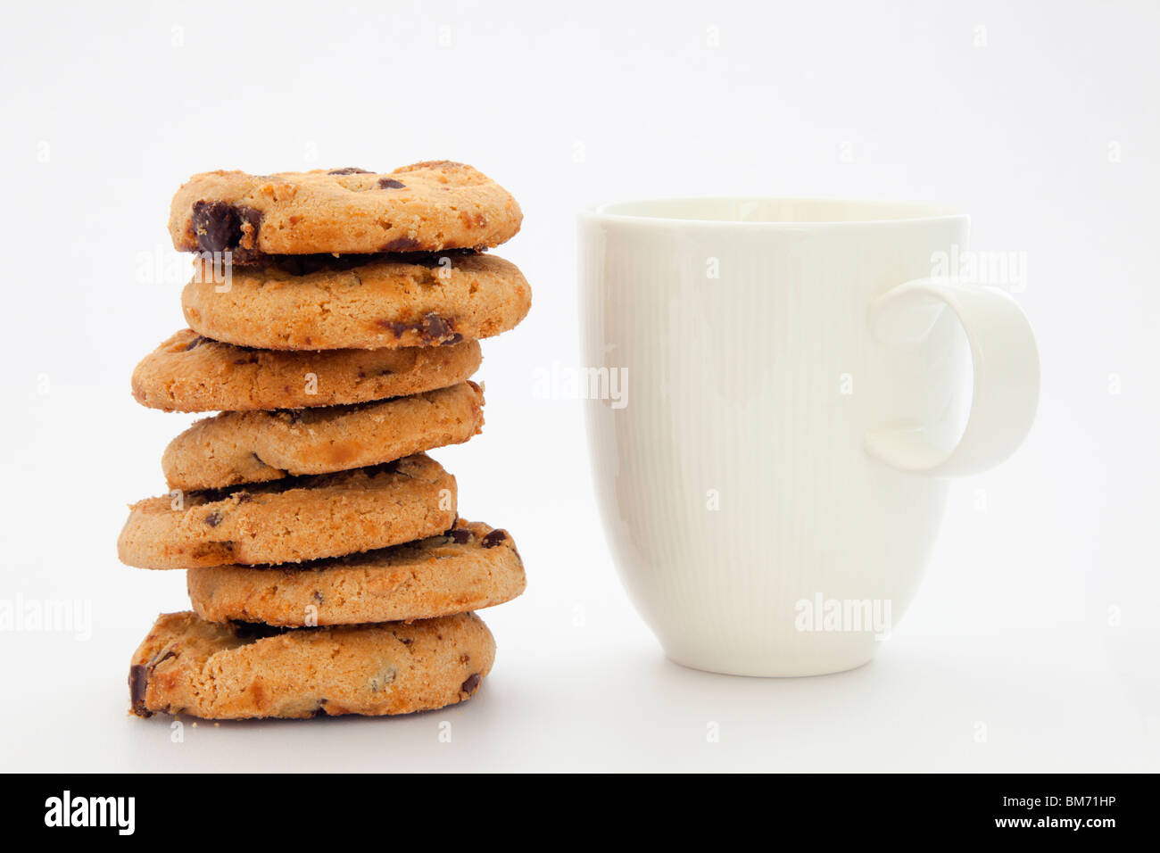Pile stack of chocolate chip biscuits and white mug of tea or coffee on a plain background. England, UK, Britain Stock Photo