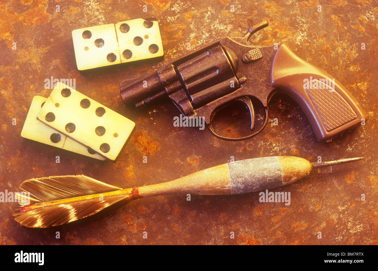 Old dartboard dart lying on rusty metal sheet in warm light with three domino pieces and toy pistol Stock Photo