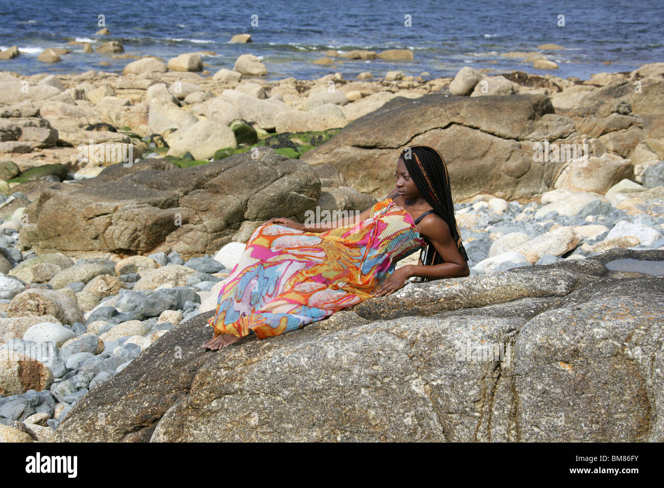 African Woman with Dreadlocks, and Wearing a Colourful Dress, Laying on Rocks by the Sea. Stock Photo